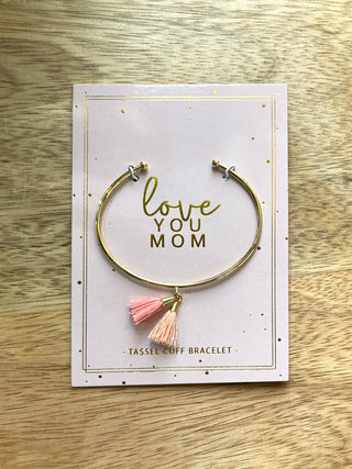 a gold cuff bracelet with double tassels in pink and peach mounted on card stock with the message love you mom