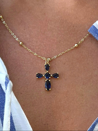 Gold necklace with a blue gemstone cross pendant on a person wearing a white and blue striped shirt.