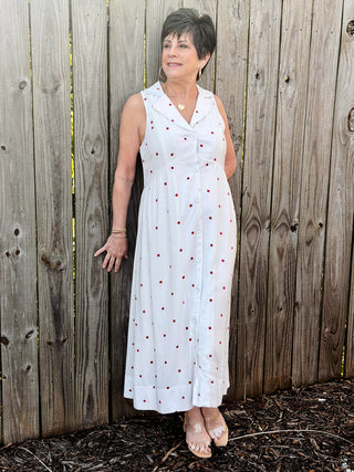 Woman wearing a white sleeveless dress with red patterns against a wooden fence.