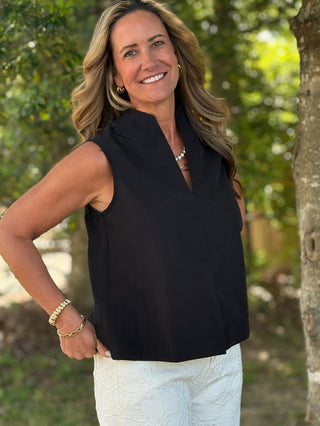 Woman wearing a black sleeveless top and white pants standing outdoors with trees in the background