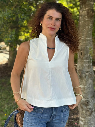 Woman wearing a white sleeveless top and blue jeans standing outdoors with greenery in the background