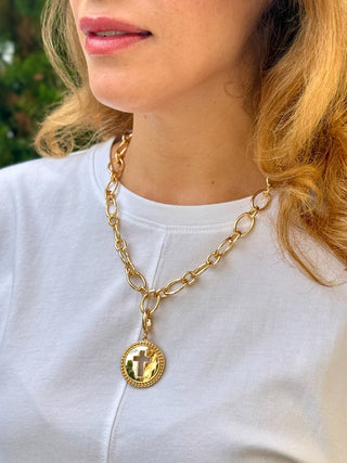a shiny round gold charm with a cross and jewels around the edge worn on a chunky gold chain necklace