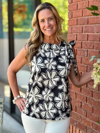 Woman wearing a black floral top and white pants standing against a brick wall.