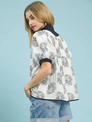 a bold abstract white blouse with a blue embroidered floral pattern worn with jean shorts