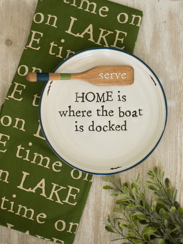 a white stoneware appetizer plate reading home is where the boat is docked with a wooden boat oar spreader reading serve