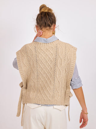 a ribbed knit sleeveless oatmeal colored sweater vest with side ties worn over a gray top