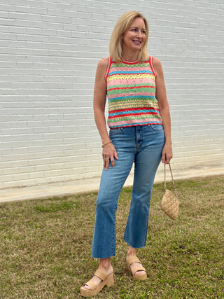 Woman wearing a colorful striped tank top and blue jeans standing in front of a white wall.