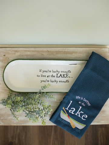 an off white stoneware tray with green trim and a navy blue waffle weave tea towel that reads life is better at the lake