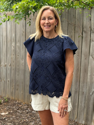 Woman wearing a navy lace top and white shorts standing outdoors with a wooden fence and greenery in the background.