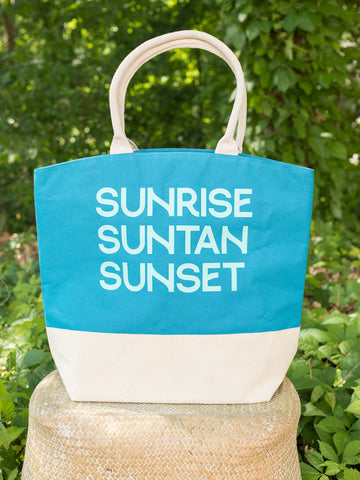 a sunrise tropic tote bag in blue jay with white sand contrast bottom and canvas handles reads sunrise suntan sunset