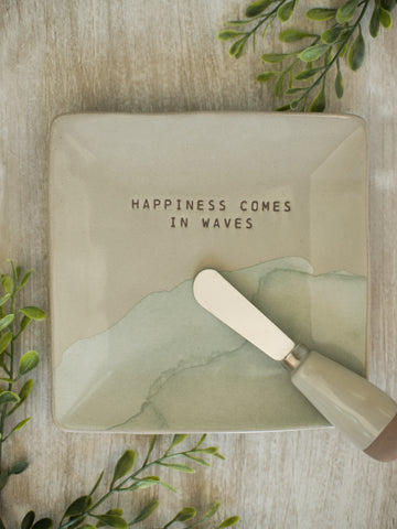 a beige square plate with sea foam green artwork reading happiness comes in waves and a matching stainless steel spreader