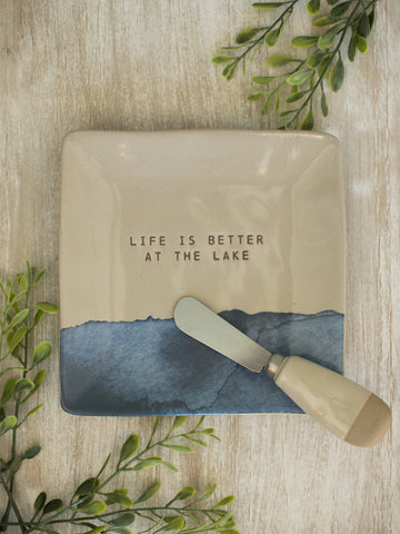 a beige square plate with dark blue artwork reading life is better at the lake and a matching stainless steel spreader