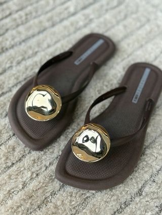 a pair of sleek brown flip flop sandals with a large gold circle at the center