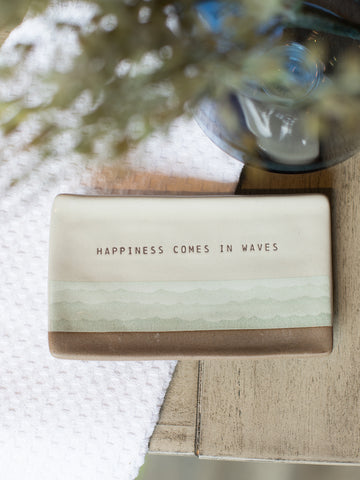 a beige rectangular spoon rest with pale green abstract wave artwork that reads happiness comes in waves