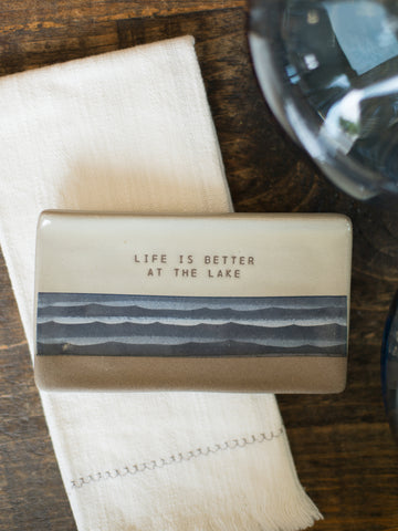 a beige rectangular spoon rest with dark blue abstract wave artwork that reads life is better at the lake