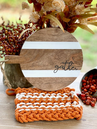 a wooden trivet that reads gather with an orange and white crochet pot holder perfect for thanksgiving host gifts