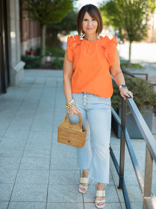 an orange ruffled sleeveless top with pleated crepe sleeves and a ruffled round neckline shown with light wash denim pants