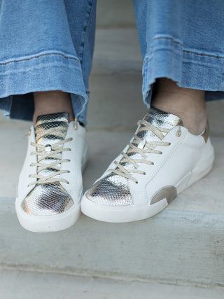 a pair of lowtop sneakers with white and gold metallics and a snake skin pattern shown with denim pants