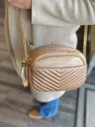 a rose gold purse made of vegan leather with gold hardware and tassel zippers shown with a beige cardigan